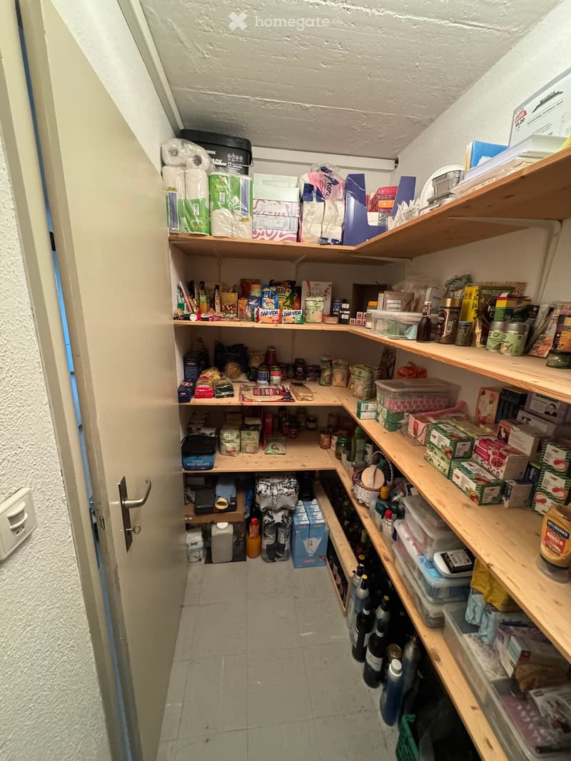A narrow pantry or storage area with wooden shelves filled with various food items, beverages, and household supplies. The space has tiled flooring and white walls.
