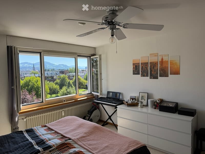 Bright bedroom with large windows offering a scenic view, featuring a bed with a colorful blanket, a white dresser, a small keyboard, and decorative items on the dresser.