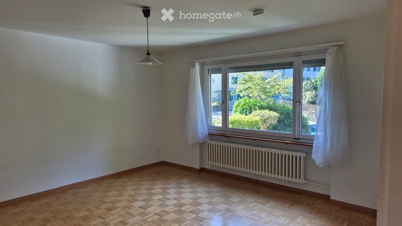 Empty room with wooden flooring, large windows with white curtains, radiator, and a ceiling light fixture.