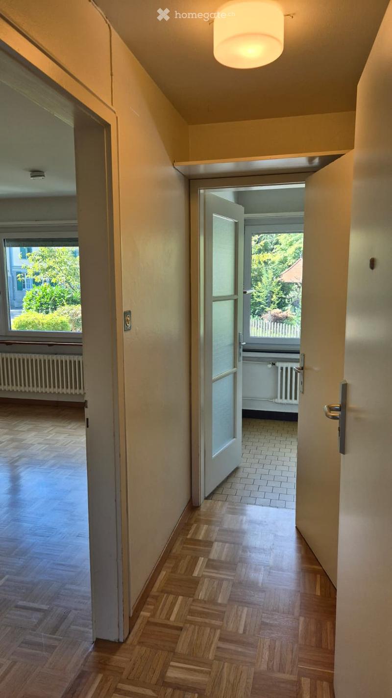 A narrow hallway with wooden flooring, light-colored walls, and a ceiling light fixture. Open doors lead to adjacent rooms, one with a view of greenery outside.