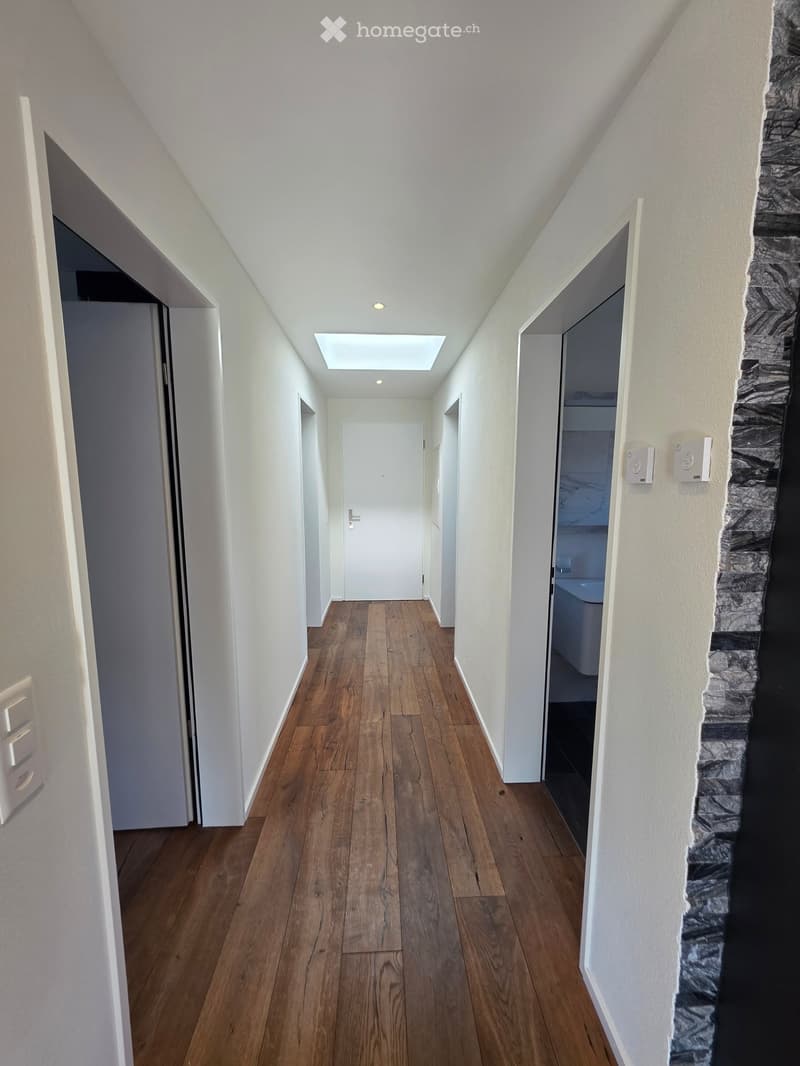 A modern hallway with wooden flooring, white walls, and recessed lighting. Features include a skylight, several doors leading to other rooms, and a stone accent wall on one side.