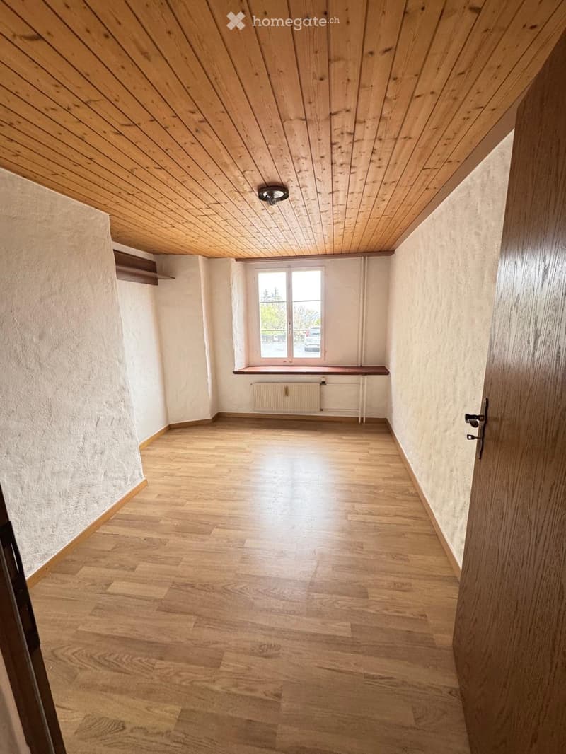 Empty room with wooden flooring, white walls, and a small window with a built-in bench and radiator below it.