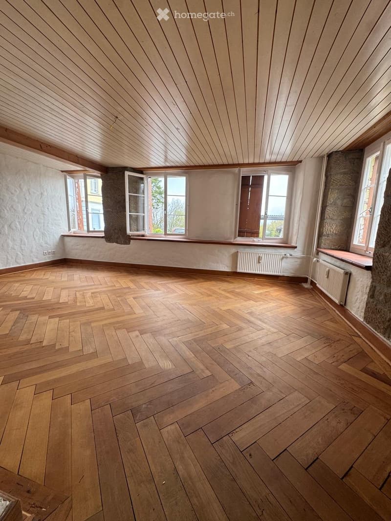 Empty room with wooden herringbone flooring, exposed wooden ceiling beams, and large windows with wooden frames. The walls are painted in a light neutral color, and there are radiators along the walls.