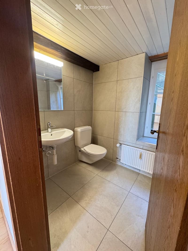 Modern bathroom with beige tiles, a sink, toilet, mirror, and a radiator. The room has a slanted ceiling and a window letting in natural light.