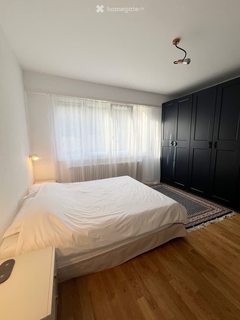 Bright bedroom with wooden flooring, a large bed with white bedding, a dark wardrobe, a nightstand, and wall-mounted lamps.