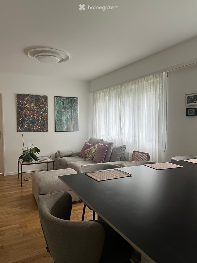 Modern living room with a grey sectional sofa, ottoman, side table with plant and books, and a dark dining table with chairs. Large windows with white curtains and abstract artwork on the walls.
