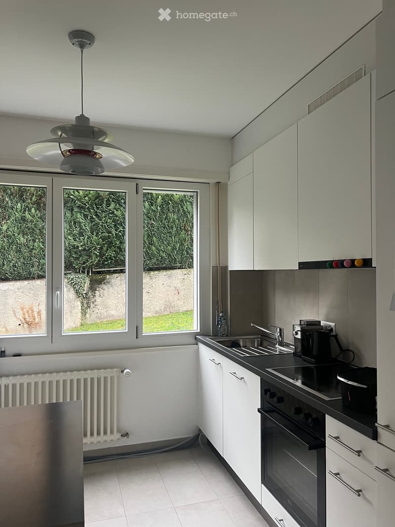 Modern kitchen with white cabinets, black countertops, stainless steel sink, oven, and coffee maker. Large windows provide natural light.