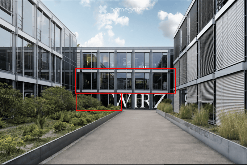 Modern multi-story building with large glass windows and sleek metallic framing, surrounded by landscaped greenery and a paved walkway.