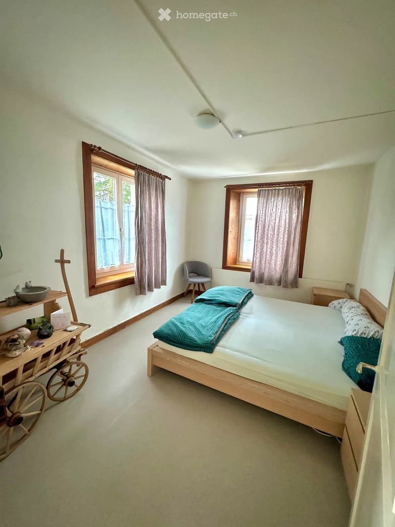 Cozy bedroom with a wooden bed, two windows with curtains, a small wooden table with decorative items, and a simple chair.