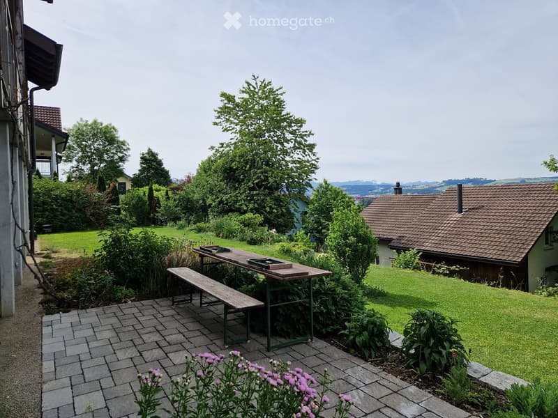 Garden area with a wooden table, bench, and surrounding greenery, including flowers and trees, with a view of a house and landscape in the background.