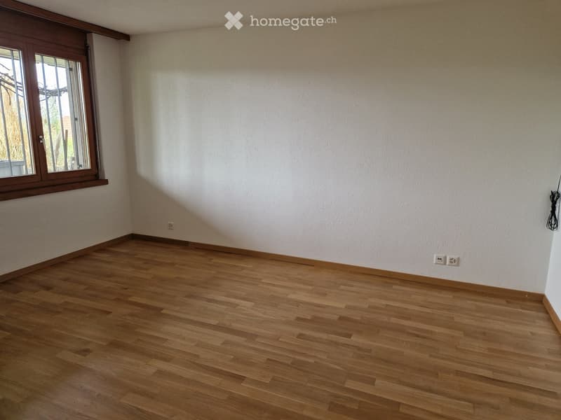 Empty room with light wooden flooring and white walls, featuring a large window with wooden frame and visible electrical outlets.