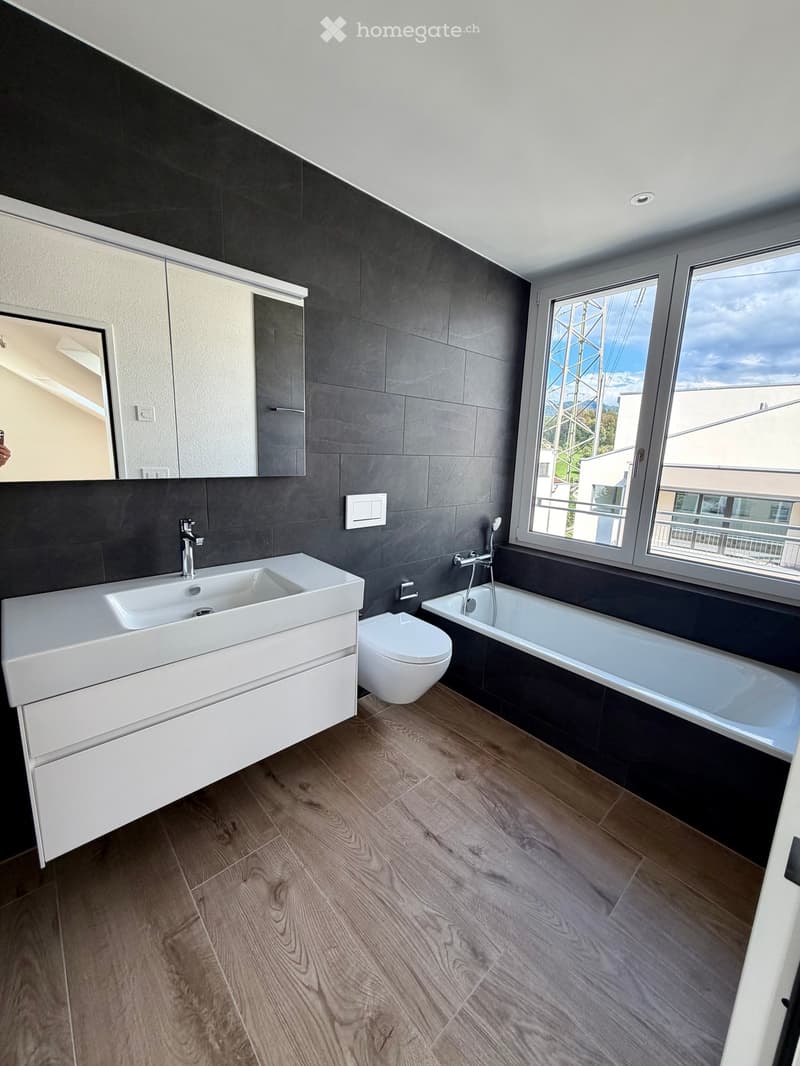 Modern bathroom with dark grey tiles, white sink, toilet, bathtub, and large windows offering a view.