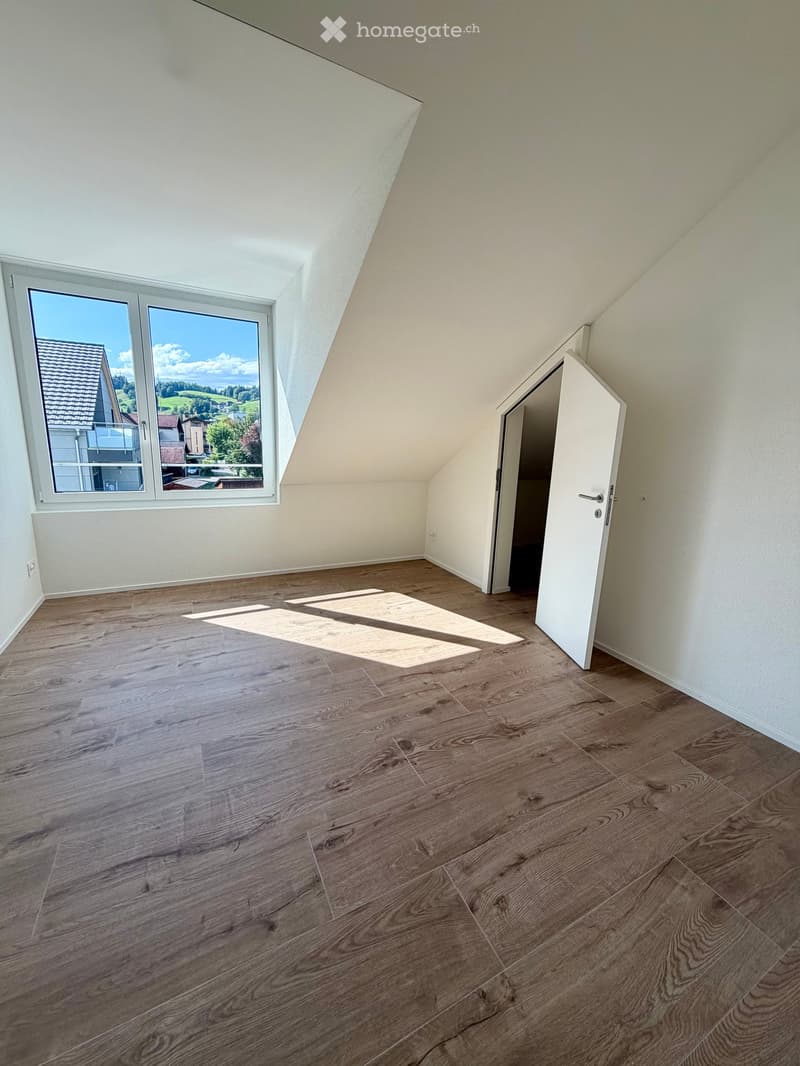 Empty attic room with wooden flooring, white walls, large window with scenic view, and an open door leading to another space.