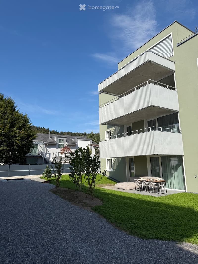 Modern two-story house with green exterior, large windows, and a balcony. Outdoor seating area with a table and chairs is visible on the ground level.