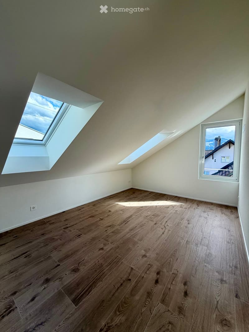 Empty attic room with wooden flooring, white walls, skylight windows, and a view of the outside through a window.