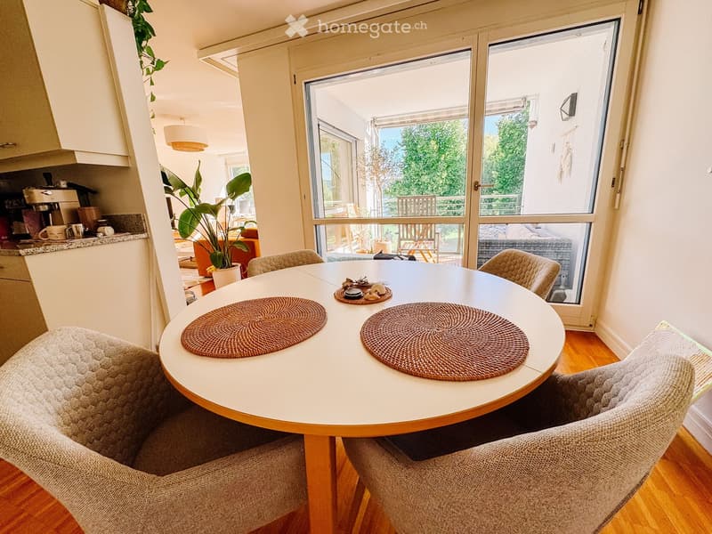 A modern dining area with a round table, four chairs, placemats, and a potted plant. The room features light-colored walls, wooden flooring, and a view of a balcony through large windows.
