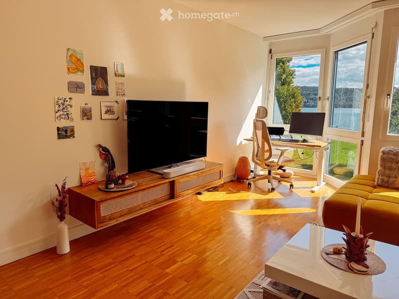 Bright living room with wooden flooring, large windows offering a scenic view, a yellow sofa, a white coffee table, a wooden TV stand, a desk with a chair, and several decorative items including vases and picture frames.