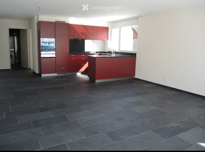 Modern kitchen with red cabinets, black countertops, and dark tiled flooring. Features a central island, sink, and oven.