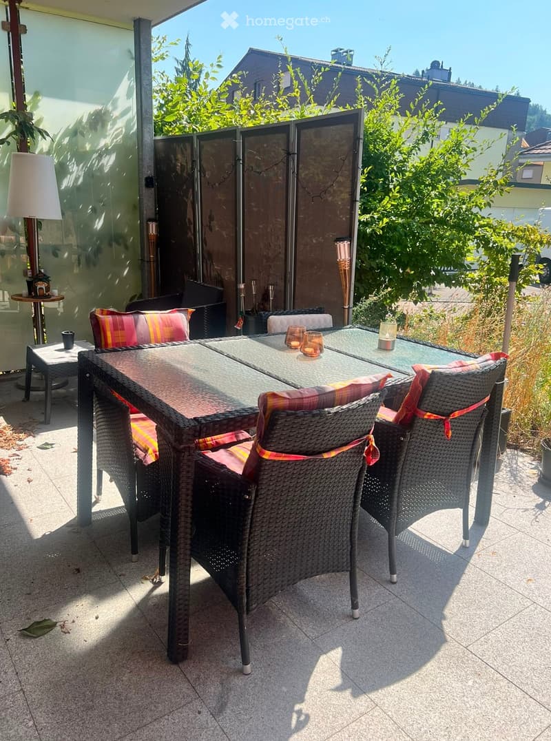 Balcony with a glass table, wicker chairs, colorful cushions, and a small side table with a lamp and cup.