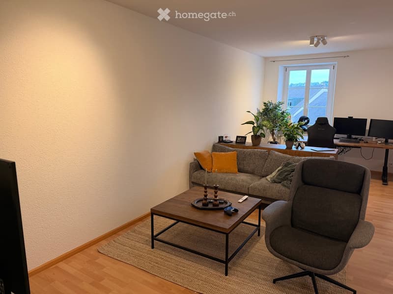 A modern living room with a grey sectional sofa, wooden coffee table, armchair, and a desk with a computer setup. The room features wooden flooring, large windows, and decorative plants.