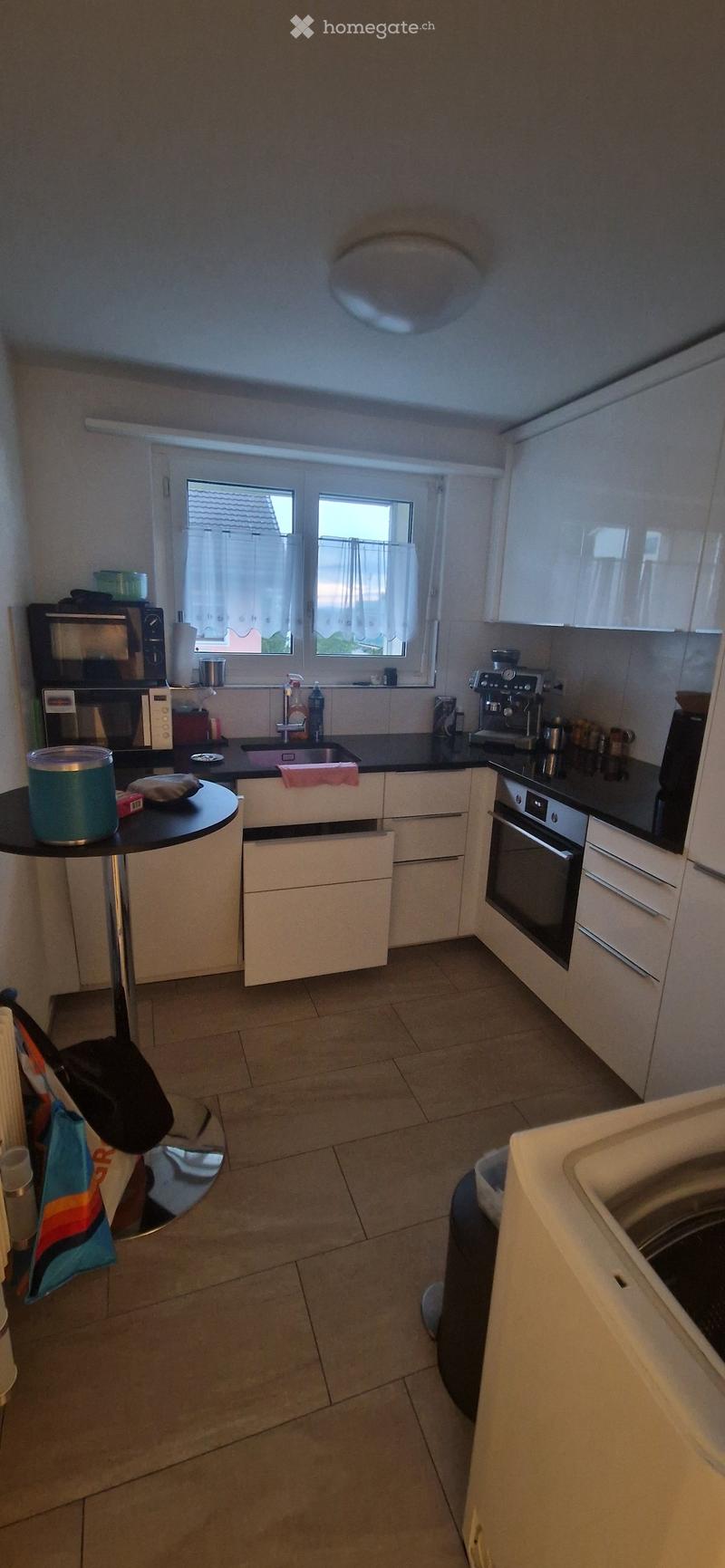 Modern kitchen with white cabinets, black countertops, and stainless steel appliances including a microwave, oven, and coffee maker. Features a sink, faucet, and a small round table with a cup and other items.