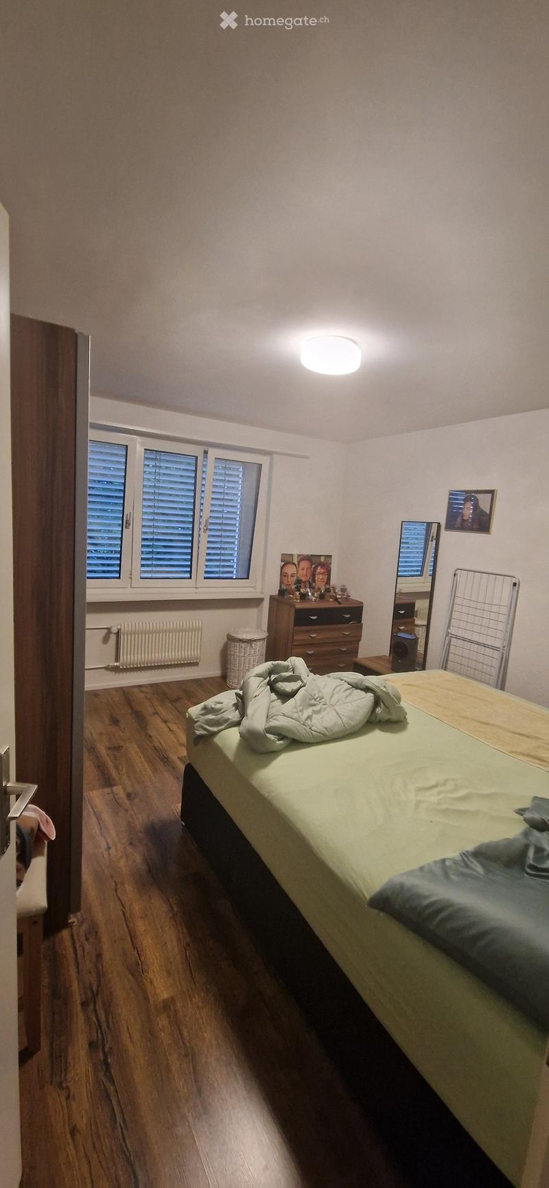 Bedroom with wooden flooring, a bed with green bedding, a nightstand with a mirror, a radiator, and a picture frame on the wall.