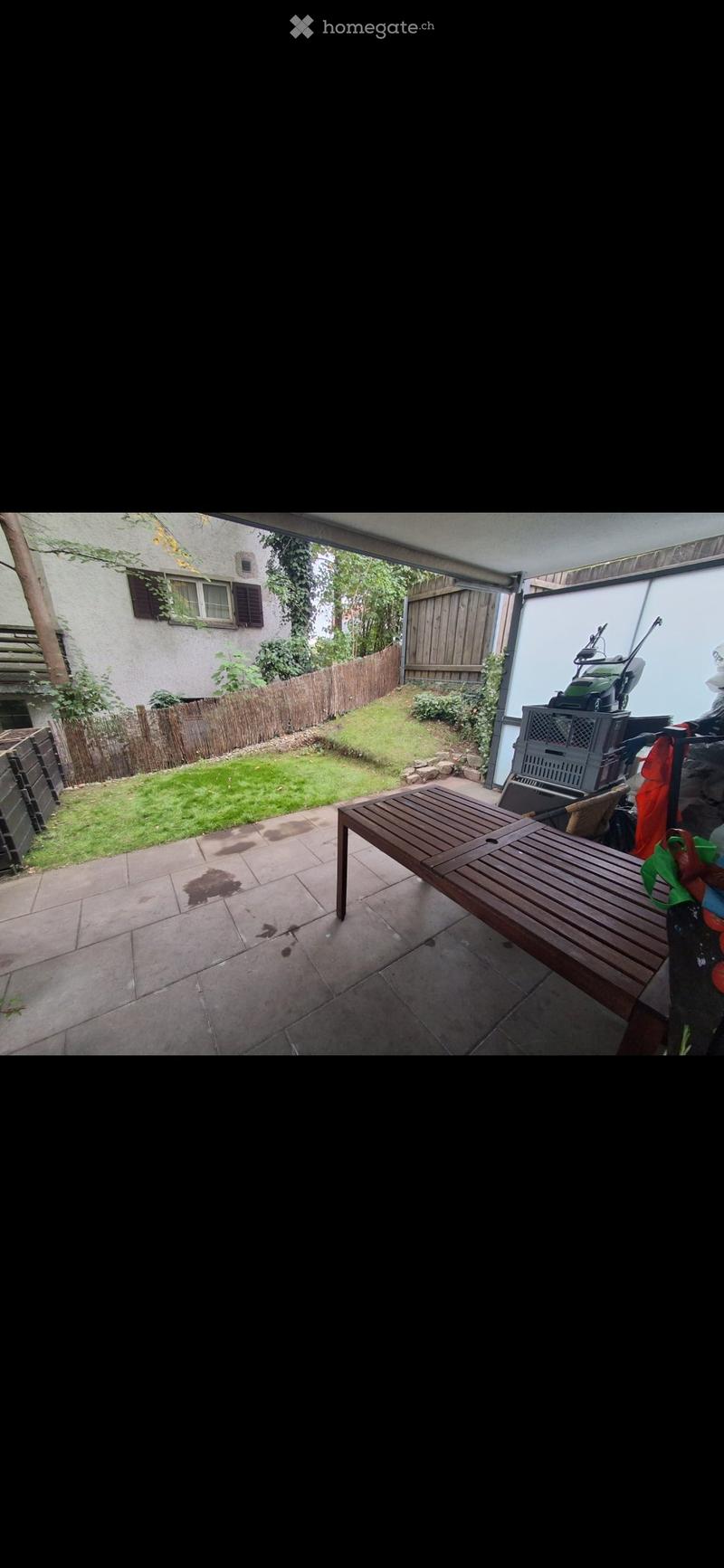 Patio area with tiled floor, wooden bench, grassy lawn, wooden fence, and neighboring house visible.