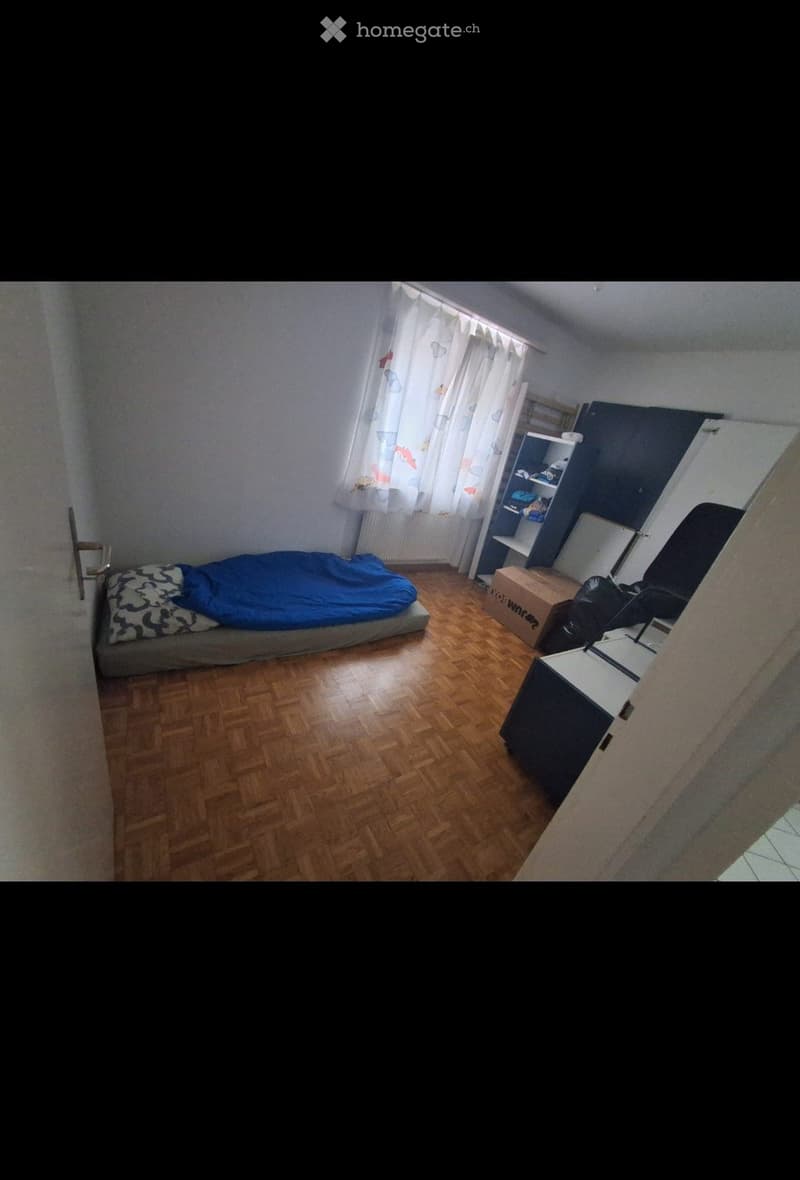 A small bedroom with wooden flooring, a bed with a blue blanket, a shelf, a chair, and a desk.