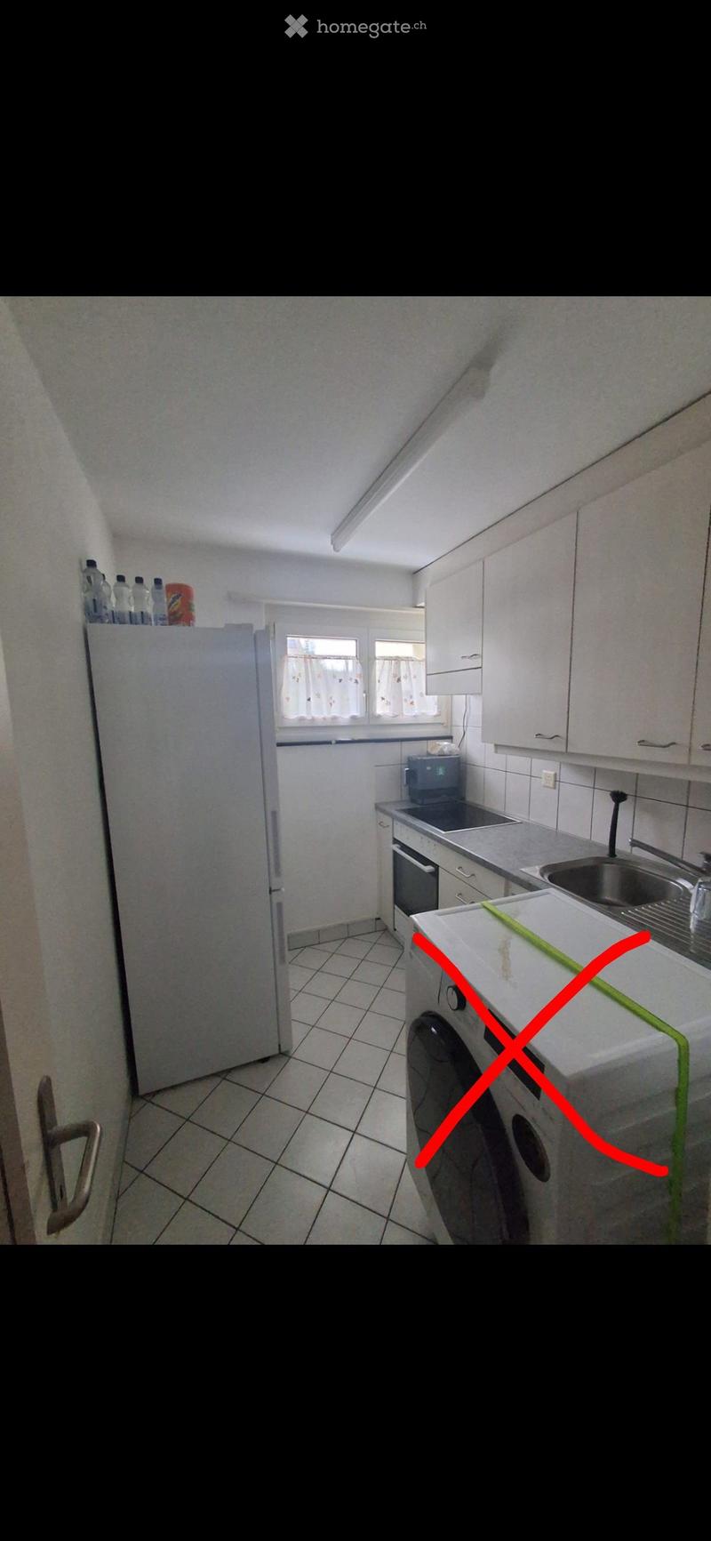 A small kitchen with white cabinets, a refrigerator, microwave, oven, sink, and washing machine. The space is well-lit with natural light from a window.