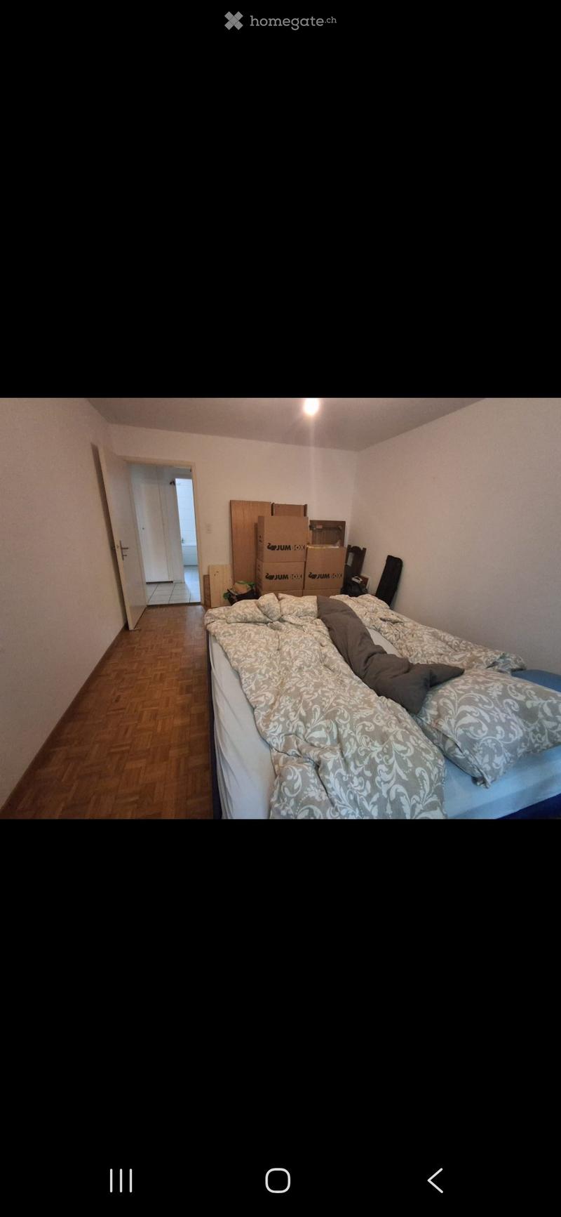 Bedroom with wooden flooring, a bed with patterned bedding, a chair, and several cardboard boxes stacked against the wall.