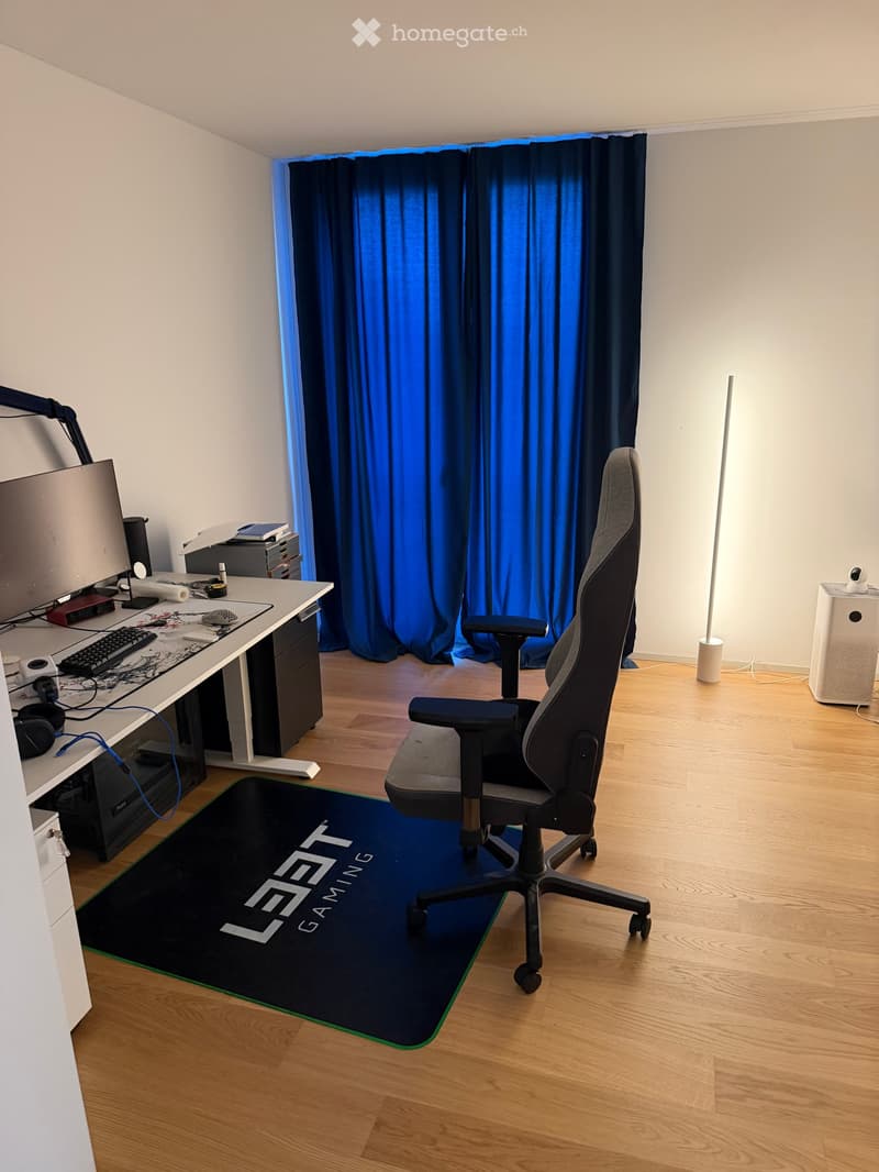 A modern, furnished room with a wooden floor, a large desk with a computer setup, a gaming chair, a blue curtain, and a standing lamp.