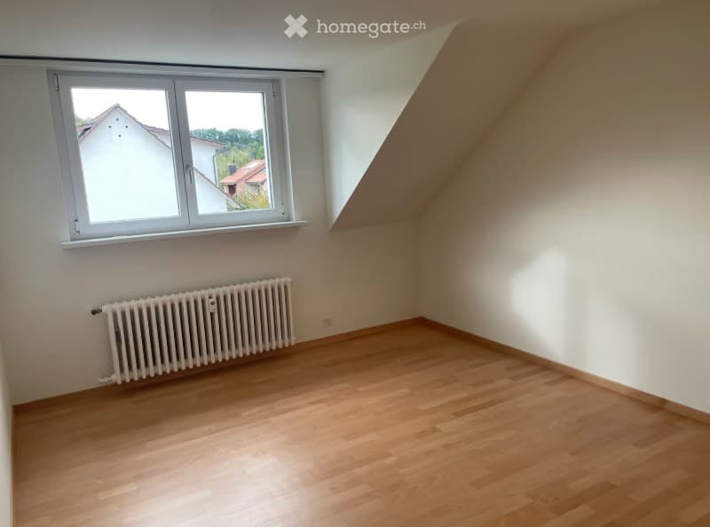 Empty attic room with wooden flooring, white walls, a single window, and a radiator. No furniture or equipment present.