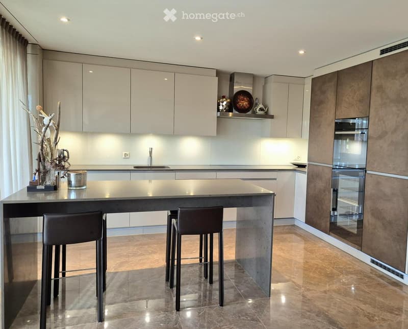 Modern kitchen with a central island, bar stools, built-in oven, sleek cabinets, and decorative items on the counter.