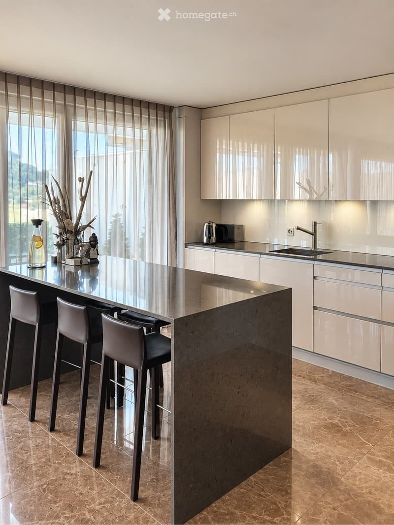 Modern kitchen with light-colored cabinets, a central island with a dark countertop, and four bar stools. The island features a vase with dried branches and a bottle on the counter. The kitchen includes a sink, microwave, and ample natural light from larg