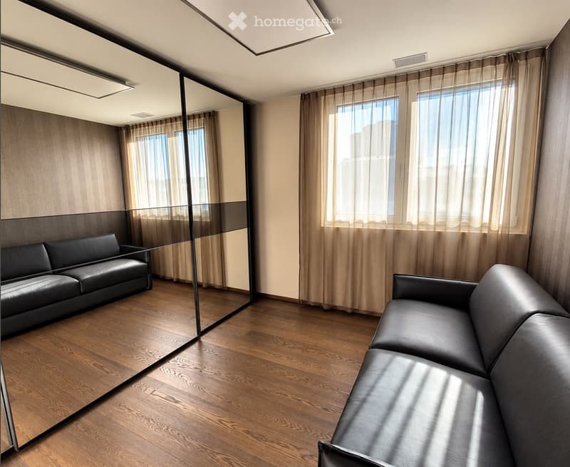 Modern living room with wooden flooring, large windows with curtains, and two black leather sofas.