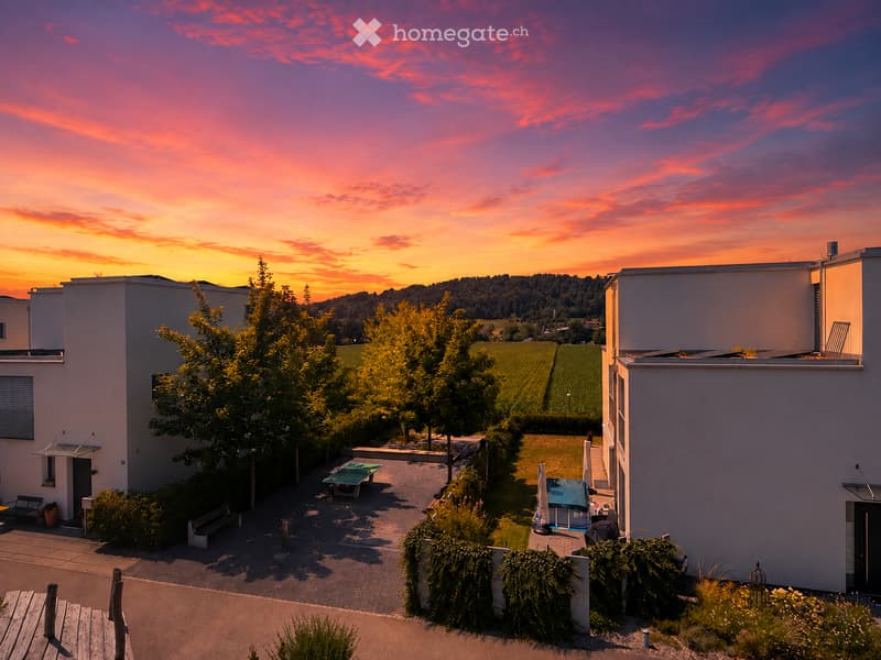 Courtyard with benches, a table, and an umbrella, surrounded by modern buildings and trees, with a scenic sunset view in the background.