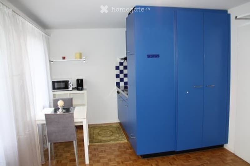 A compact, modern room with a dining table and chair, a microwave, a tall blue wardrobe, and a small shelf with decorative items.