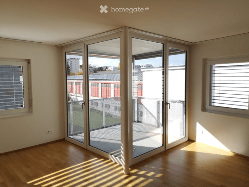 Empty room with wooden flooring, large glass sliding doors leading to a balcony, and three windows with blinds.