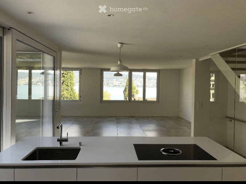 Modern, minimalist kitchen with white countertops, built-in sink, induction cooktop, and large windows overlooking a scenic lake view.