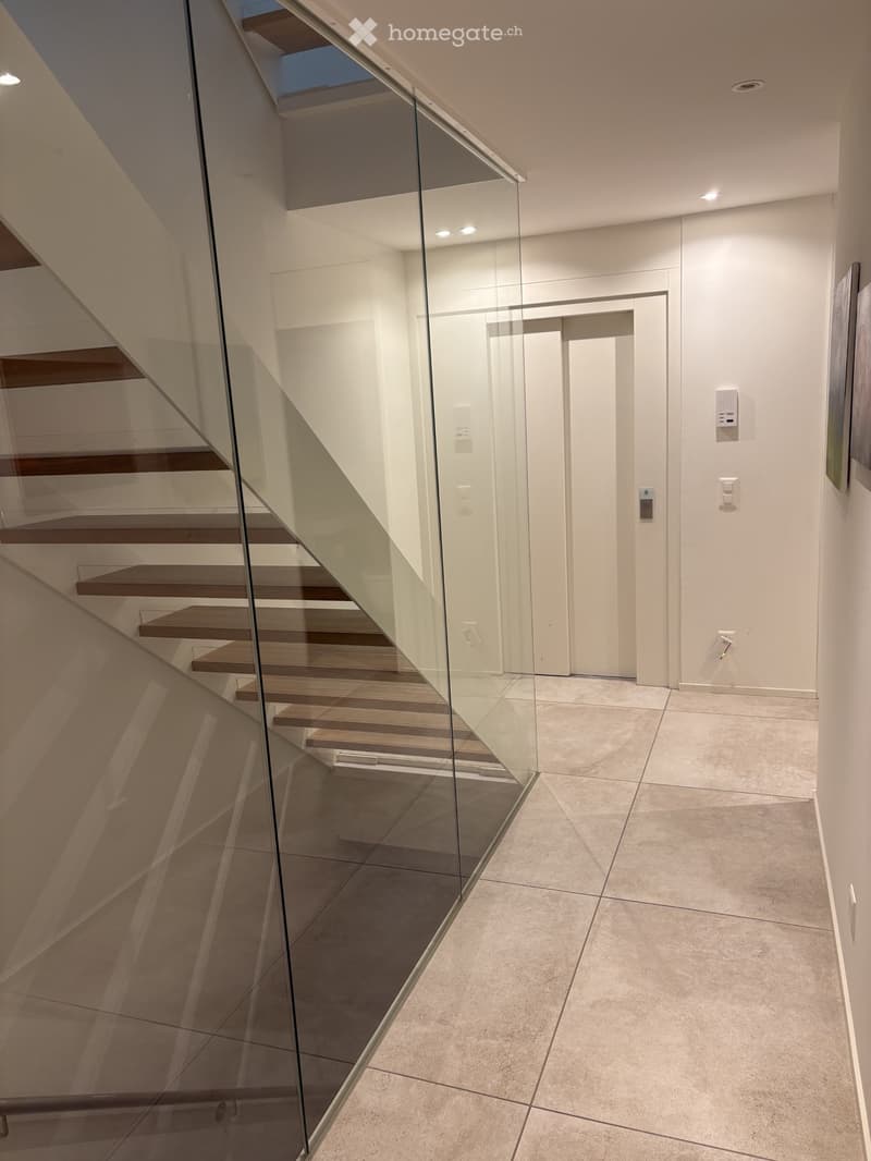 Modern hallway with a glass-walled staircase featuring wooden steps and a sleek metal railing, leading to an upper level.