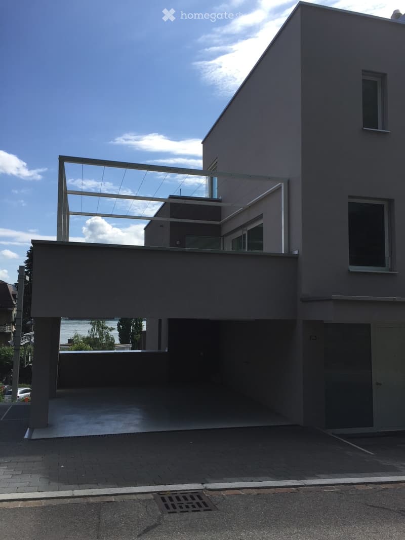 Modern two-story building with a flat roof, large windows, and a covered parking area. The structure features a minimalist design with clean lines and a neutral color palette.