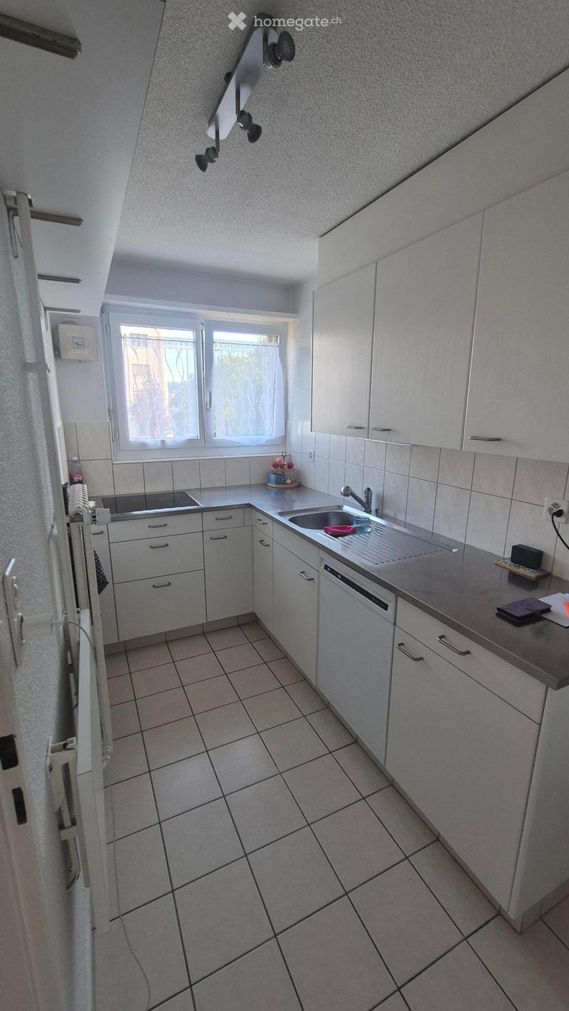 Modern kitchen with white cabinets, tiled floor, and stainless steel sink. Features a window with blinds and a small countertop display with decorative items.