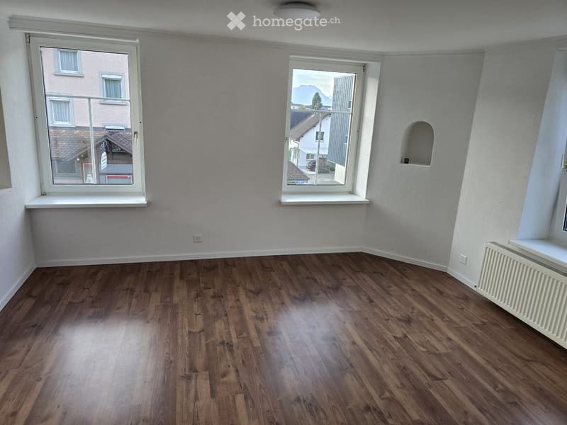 Empty room with wooden flooring, white walls, and large windows offering a view of the outside. The room features a radiator on one wall and a small niche in the corner.