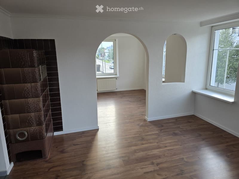 Empty room with wooden flooring, white walls, large windows, and an arched doorway leading to another space.