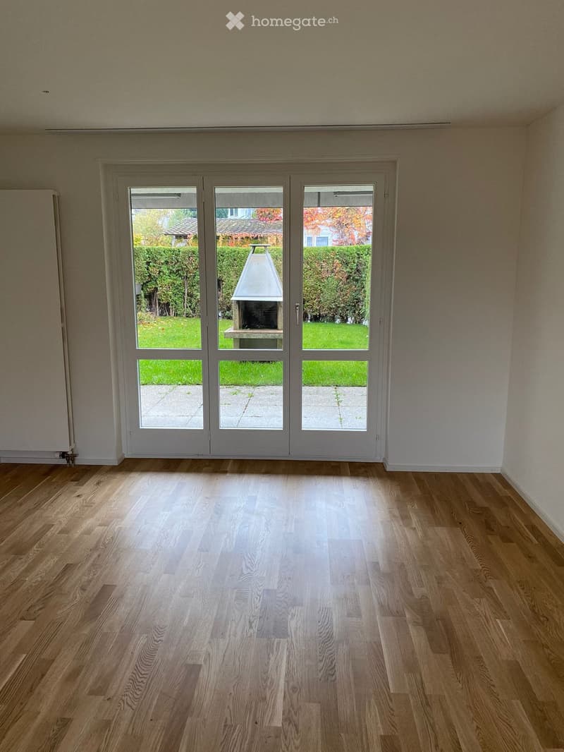 Empty room with wooden flooring and large glass doors leading to an outdoor area with a barbecue and garden.
