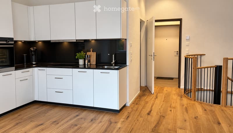 Modern kitchen with white cabinets, black countertops, and stainless steel appliances. A small plant and coffee maker are on the counter. The adjacent area shows a wooden staircase with a railing.