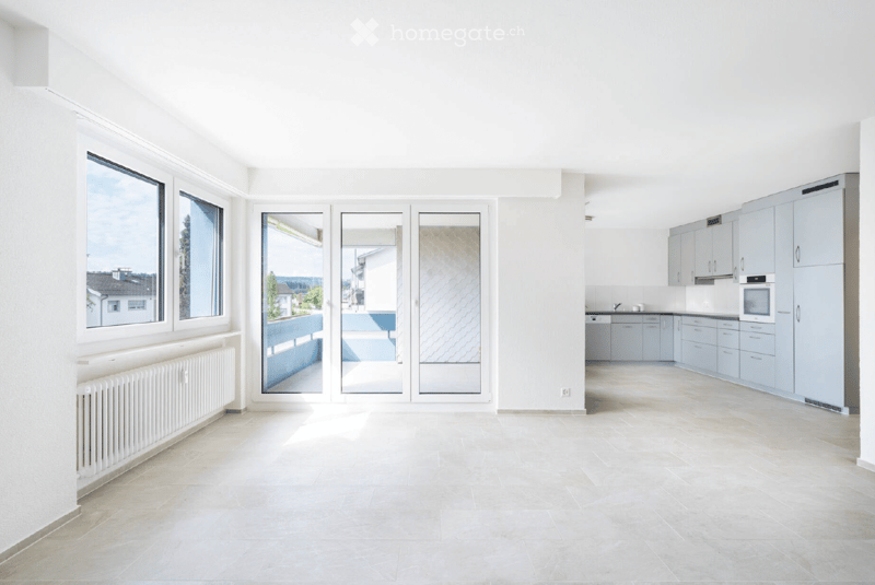 Bright, open room with large windows and sliding glass doors leading to a balcony. Light grey cabinets and appliances are visible in the adjacent kitchen area.