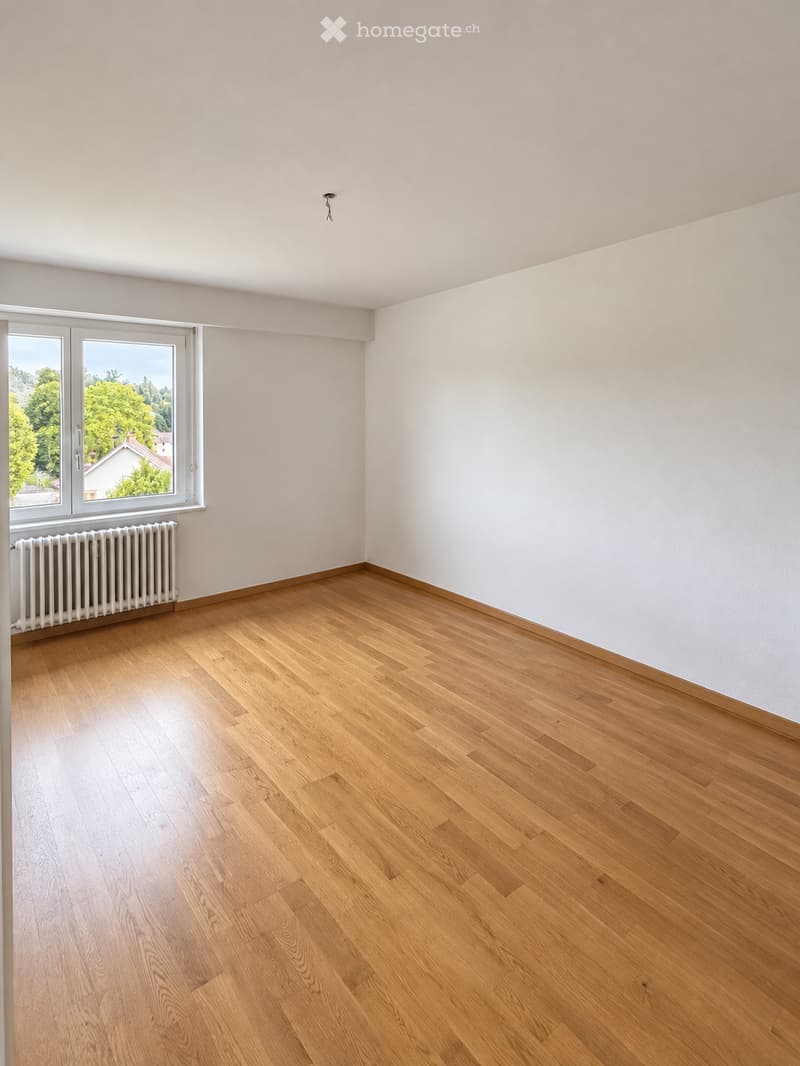 Empty room with wooden flooring, white walls, and a large window with a view of greenery and a house outside. A radiator is visible below the window.