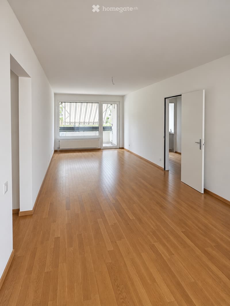 Empty room with wooden flooring, white walls, and large windows with blinds. There is a radiator under one window and a door leading to another room.