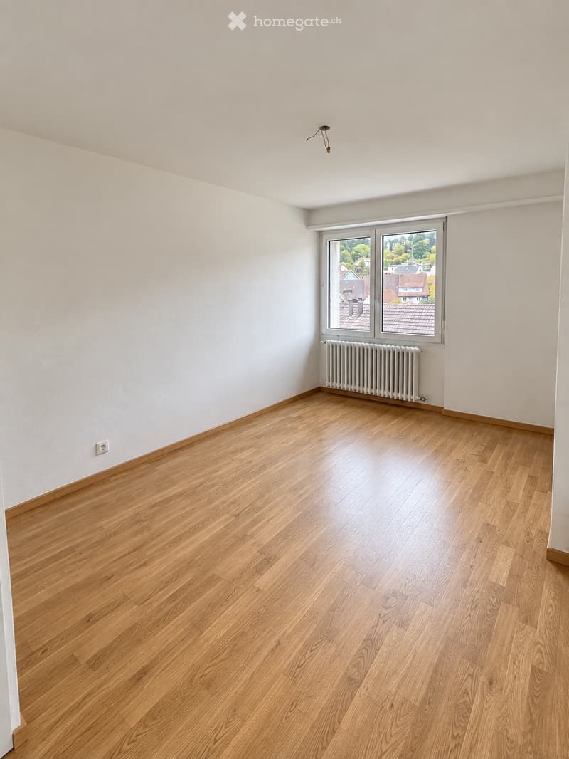 Empty room with light wooden flooring, white walls, and a large window with a radiator below it.